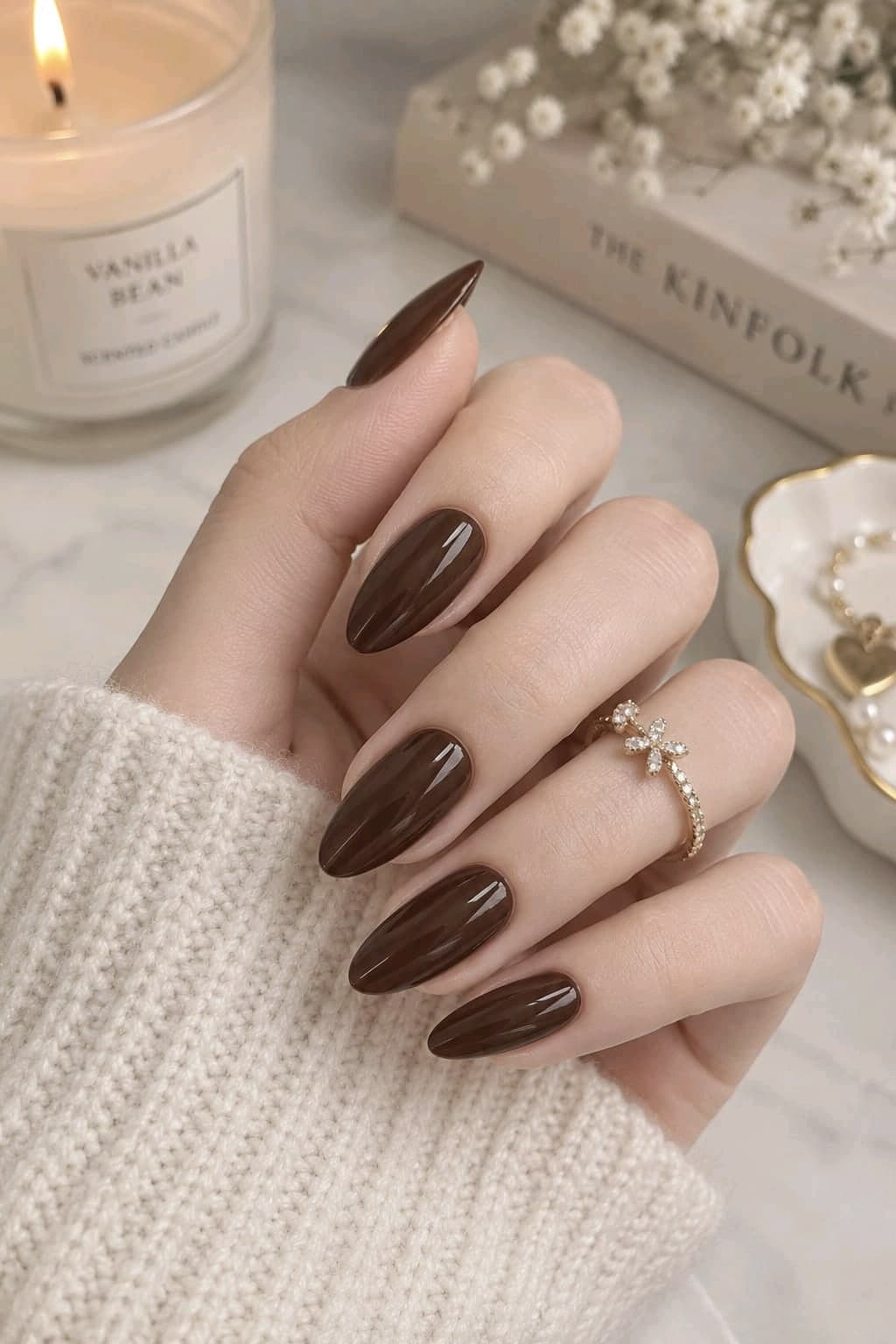 Glossy chocolate brown almond nails with a gold flower ring and a cream knit sweater