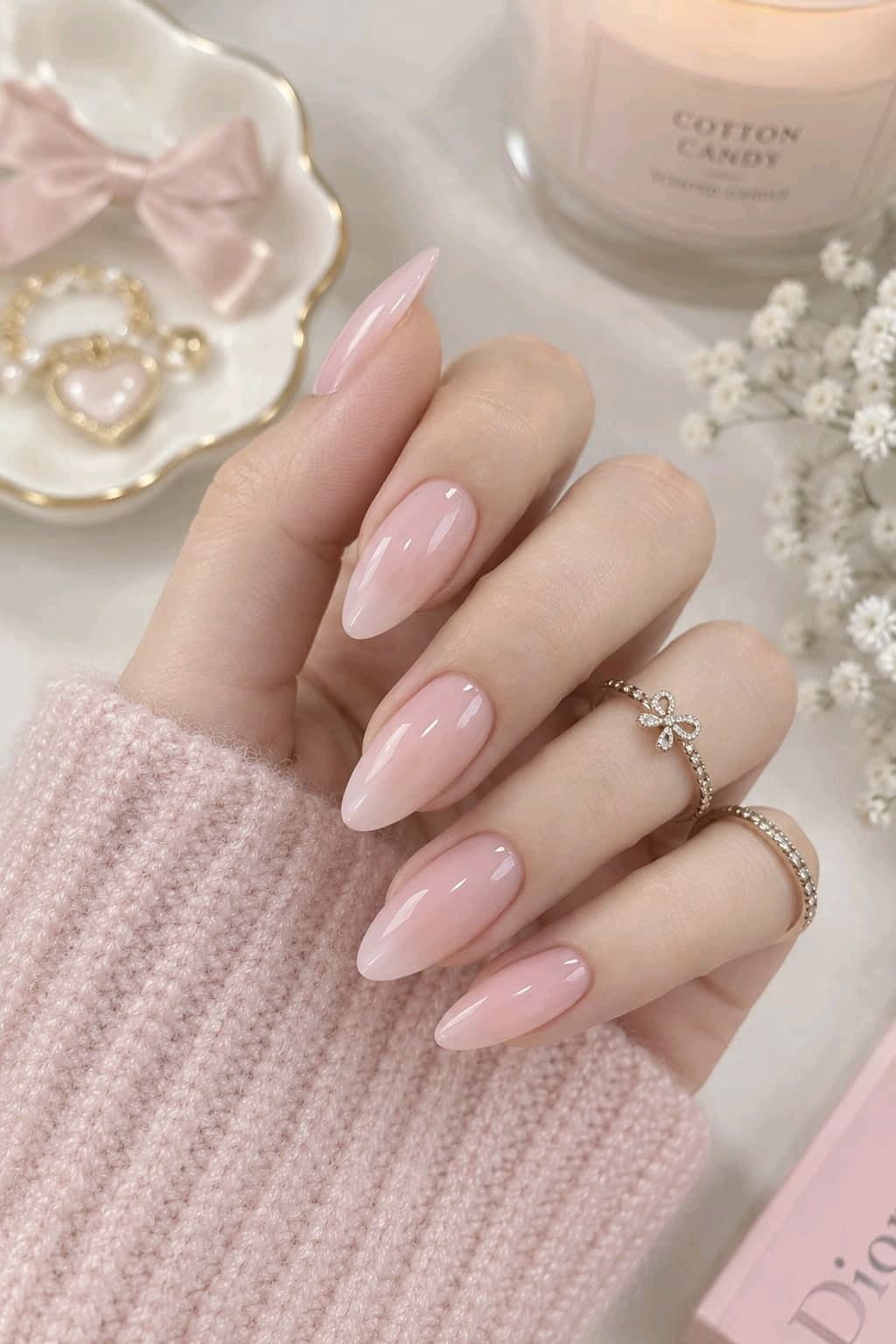 Sheer milky pink almond nails with a gold bow ring and a pink knit sweater