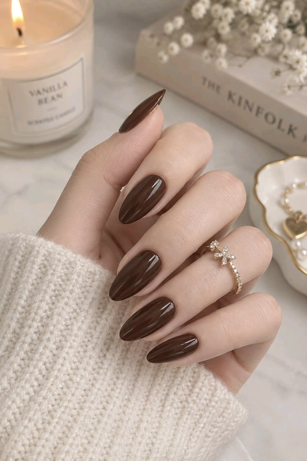 Glossy chocolate brown almond nails with a gold flower ring and a cream knit sweater